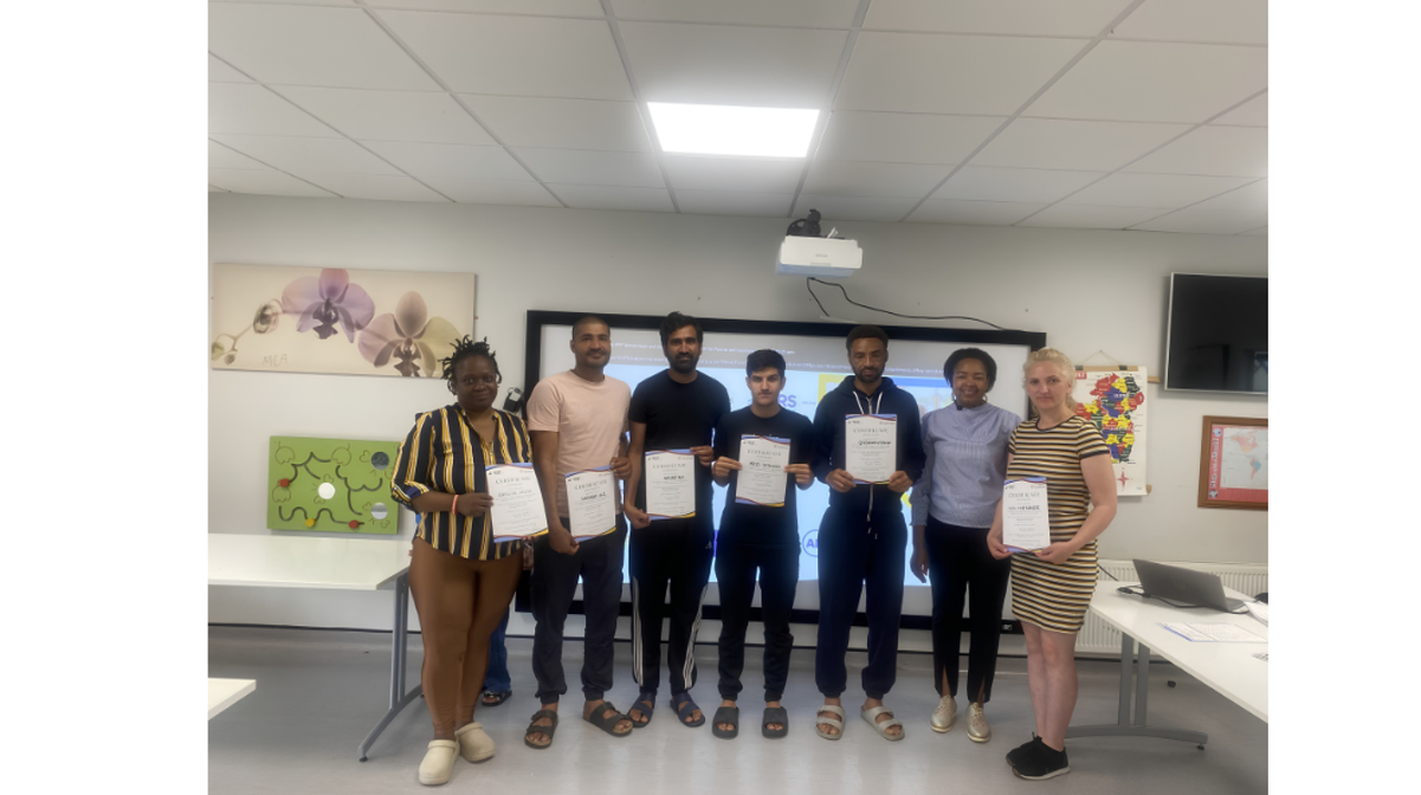 Seven people standing in a classroom, holding certificates, with a projector screen and wall art in the background.